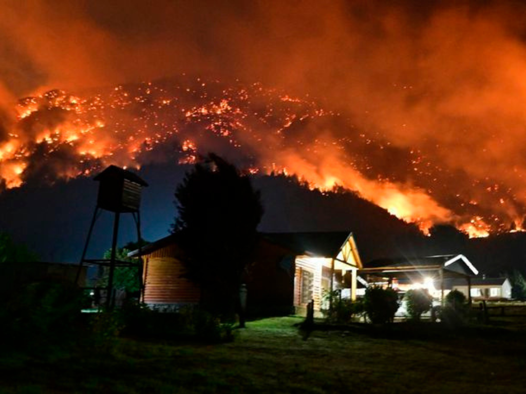 Cabaña de madera en la zona cordillerana con fuego y humo avanzando sobre las montañas durante la emergencia en Los Alerces.