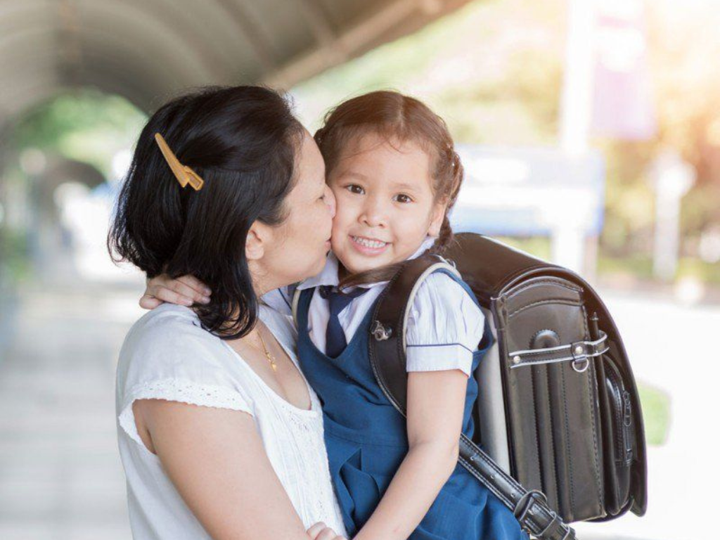 Madre despidiendo a su hija con uniforme y mochila para el inicio de clases, representando la Ayuda Escolar ANSES.