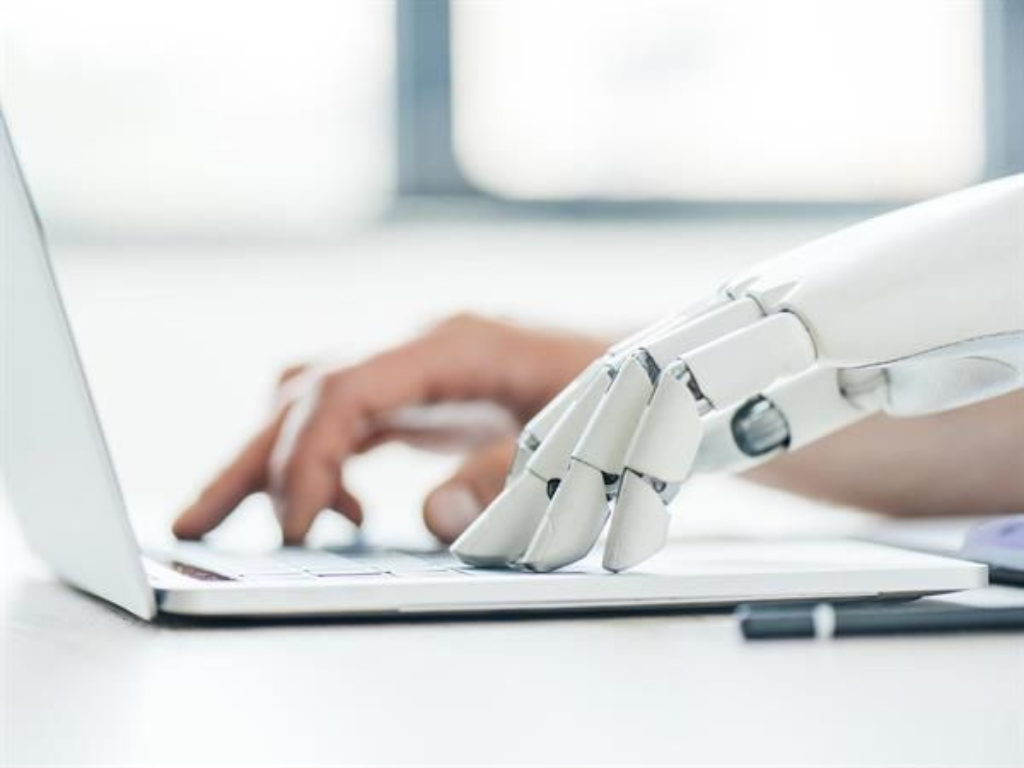 Primer plano de una mano robótica y una mano humana desenfocada sobre el teclado de una laptop, representando el impacto de la IA y mercado laboral.